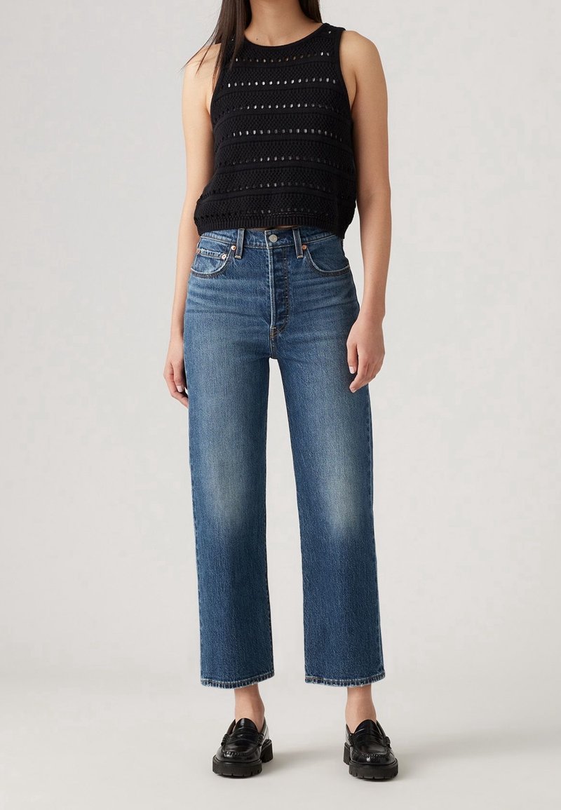 Femme portant un haut en crochet noir sans manches, un jean droit bleu taille haute et des mocassins noirs, debout devant un fond uni.