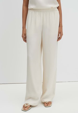 Vrouw die losse, hoog getailleerde brede crèmebroek draagt in combinatie met crème teenslippers, staand tegen een effen achtergrond.