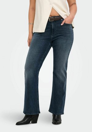 Femme portant un jean droit bleu foncé, des bottines noires et un top beige sans manches, main dans la poche, debout sur fond blanc.
