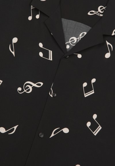 Zwart T-shirt met korte mouwen, gemaakt van stof met witte muzieknoten en symbolen in verschillende groottes over het oppervlak verspreid. Dichtknopen aan de voorkant.