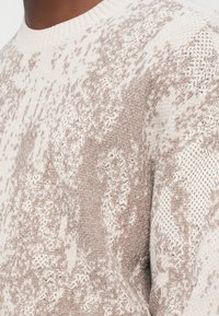 Μπεζ πλούσιο πουλόβερ με σχέδιο μικτής υφής και πιτσιλιές ανοιχτού καφέ, στρογγυλή λαιμόκοψη και ριμπ ύφανση στα μανίκια και στο τελείωμα.
