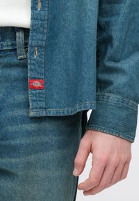 Ανοιχτό μπλε πουκάμισο από τζιν με κόκκινο ταμπελάκι Dickies κοντά στο τελείωμα, συνδυασμένο με τζιν παντελόνι σε ίδιο χρώμα. Ορατή ραφή και υφή υφάσματος.