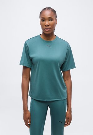 Vrouw met gevlochten haar, gekleed in een teal Puma T-shirt met korte mouwen en bijpassende legging, staand tegen een effen lichte achtergrond.