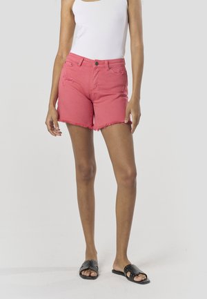 Ružové rifľové šortky s rozstrapateným okrajom, zdobené opotrebovanými detailmi, doplnené čiernymi šmykľavými sandálmi. Modelka má na sebe biele tielko.