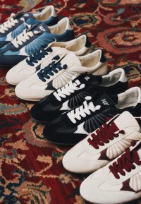 Cinq paires de baskets en bleu, blanc, noir et bordeaux avec des lacets contrastants, disposées en rangée sur un tapis rouge à motifs.