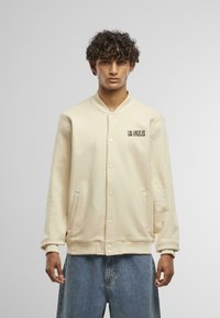 Svetlobéžová varsity bunda so zacvakávacími gomb�íkmi, ribbovanými manžetami a spodkom. Na ľavej hrudi má potlač "LOS ANGELES" v čiernej Farbe.