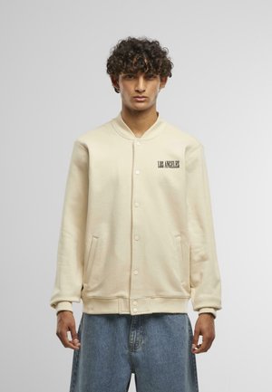 Šviesiai kreminės spalvos sportinis švarkas su snap mygtukais, raukšlėtais rankogaliais ir apačia. Kairėje krūtinės pusėje juodai spausdinta "LOS ANGELES".