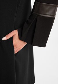 Schwarzes Kleid mit glatter Textur, das an den Ärmeln einen Lederakzent aufweist. Die Hand ist in einer Tasche an der Seitennaht positioniert.