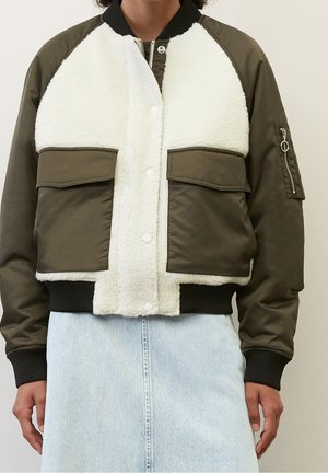 Chaqueta bomber sherpa marrón y blanca con grandes bolsillos delanteros, puños negros y una falda vaquera azul claro visible debajo de la chaqueta.