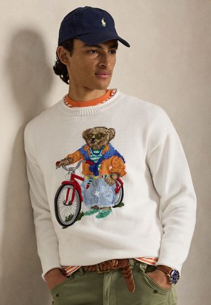 Νεαρός άνδρας που φοράει μπλε σκούφο, λευκό πουλόβερ με αρκούδα πάνω σε κόκκινο ποδήλατο, ριγέ πορτοκαλί πουκάμισο, πράσινο παντελόνι και ρολόι, στέκεται μπροστά σε απλή τοίχο.