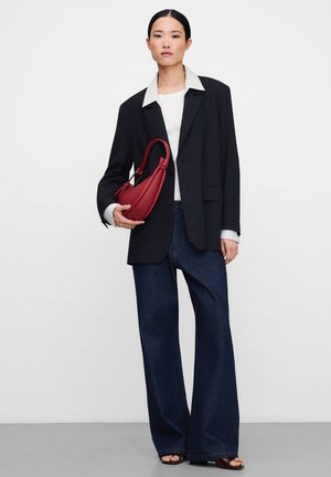 Vrouw met donker haar, een zwart colbert, wit overhemd en donkerblauwe wijde jeans, die een rode handtas vasthoudt tegen een effen witte achtergrond.