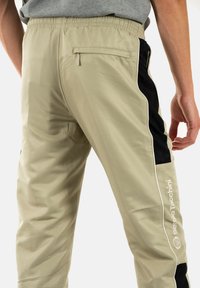 Pantalones beige con un acabado texturizado, que presentan paneles laterales negros, cintura elástica y un bolsillo trasero con cremallera. Detalle del logo en blanco.