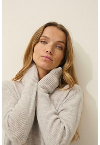 Maglione grigio chiaro a coste con colletto alto, caratterizzato da maniche lunghe e una texture morbida. Il modello posa con le mani sul colletto.