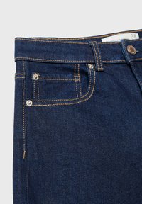 Jeans en denim bleu foncé avec des coutures orange, présentant une poche avant et des rivets en métal, soigneusement pliés à la taille avec une étiquette visible.