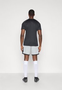 Μαύρο αθλητικό μπλουζάκι με κοντά μανίκια, συνδυασμένο με ανοιχτό γκρίζες βερμούδες με μαύρη περιφέρεια. Λευκές γόνατες κάλτσες και μαύρα παπούτσια ποδοσφαίρου.