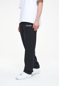 Svarta denimjeans med "PEGADOR" broderat i vitt, i kombination med en vit t-shirt och vita sneakers, vilket ger en avslappnad passform.