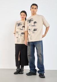 Camisetas de gran tamaño en color beige con un estampado abstracto oscuro, combinadas con una falda negra con una abertura lateral y jeans azules. Calzado negro completa el look.