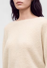 Femme aux cheveux foncés portant un pull en tricot texturé couleur crème avec un large col rond sur un fond uni.