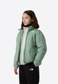 Lysegrøn vanddunjakke med lynlåslukning foran og hætte, med quiltet tekstur og logo på venstre skulder. Båret over et hvidt lag.