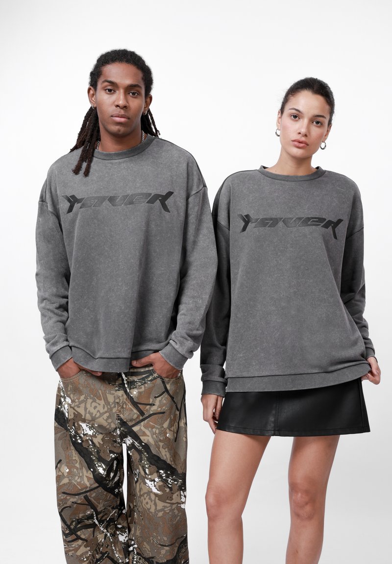 Felpe grigie oversize con un logo audace, abbinate a pantaloni mimetici a fantasia e a una gonna di pelle nera. Stile urbano casual.