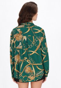 Cămașă verde cu model și accente aurii, cu lanțuri, forme sferice și ciucuri. Material moale, cu mâneci lungi și croială lejeră.