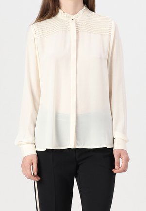 Femme portant un chemisier crème à manches longues avec des détails plissés sur la poitrine, col boutonné, associé à un pantalon noir.