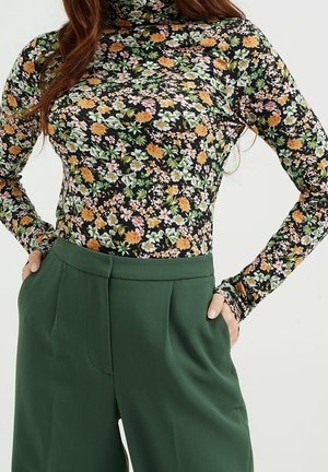 Femme portant un pantalon taille haute vert foncé et un haut noir à manches longues avec des fleurs orange, blanches et vertes, mains dans les poches.