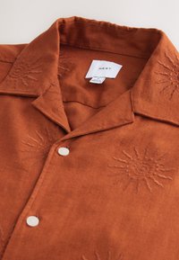 Short-sleeve, burnt orange shirt made of textured fabric, featuring embroidered sun designs and white buttons. Label visible inside the collar.