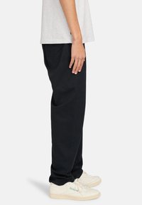 Zwarte casual broek van katoen, met een rechte snit, en gecombineerd met witte Reebok sneakers. Het model staat zijwaarts gedraaid.
