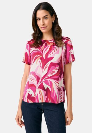 Short-sleeve blouse in pink with leaf patterns, featuring a round neckline and soft, smooth fabric. Paired with dark denim.