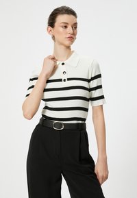 Kurzarm-Poloshirt aus geripptem weißen Stoff mit schwarzen horizontalen Streifen; verfügt über einen Kragen und eine Knopfleiste mit drei Knöpfen. Schwarze Hose und Gürtel.
