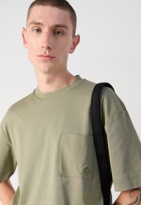 Olive spalvos trumpas rankovės marškinėliai, pagaminti iš minkštos medvilnės, turi krūtinės kišenę su siuvinėtu logotipu ir apvalų iškirpimą.