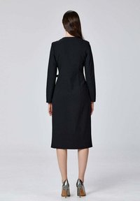 Robe noire à manches longues, ajustée, courte au genou, texture lisse et encolure ronde, associée à des chaussures à talons hauts argentées.