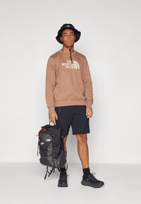 Felpa marrone con logo, cappello a secchiello nero, zaino nero con dettagli rossi, pantaloni corti neri e scarpe scure. Presenta un design semplice e funzionale.