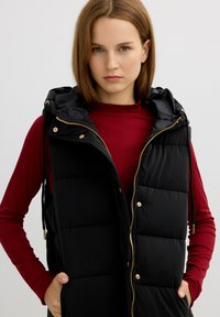 Chaleco acolchado negro con cierre de cremallera, capucha con cordón y detalles en tono dorado. Llevado sobre una camiseta de manga larga roja, que presenta un diseño elegante.