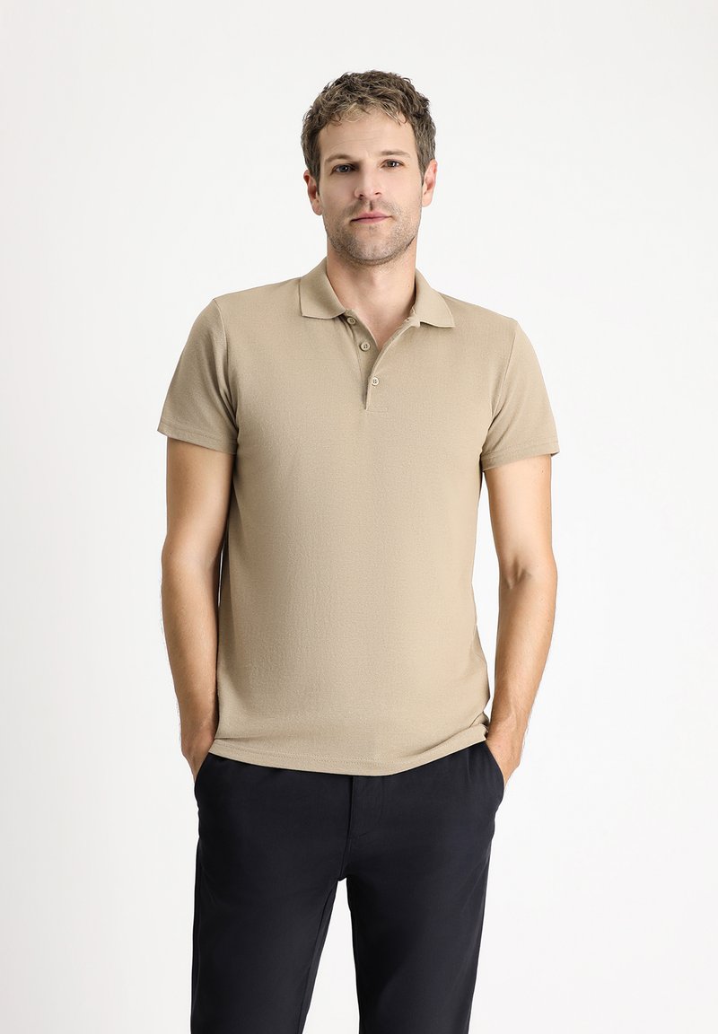 Beżowa koszulka polo z teksturowanej tkaniny, z kołnierzykiem i trzema guzikami, w połączeniu z ciemnymi spodniami, ręce w kieszeniach.