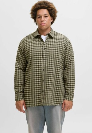 Ung mann med krøllete hår som har på seg en grønn og beige rutete skjorte med knapper og lyseblå jeans, stående foran en ensfarget hvit bakgrunn.