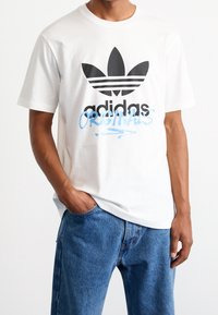 Bílá bavlněná tričko s černým logem Adidas a nápisem "ORIGINALS" ve světle modrém písmu, doplněné modrými denimovými džíny.