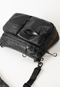 Borsa nera in tessuto dal design texturizzato, dotata di numerose tasche con zip e un'apertura frontale. Presenta spallacci regolabili e ganci metallici. Forma rettangolare.
