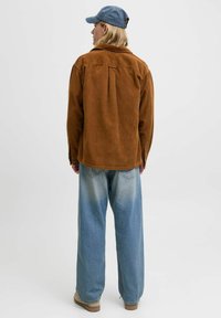Braune Cord-Hemd mit lockerer Passform, kombiniert mit hellblauen Weitbeinen-Jeans und beigen Schuhen, von hinten betrachtet. Blaue Mütze auf dem Kopf.