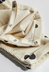 Crème-kleurige corduroy stof met zwarte stippen, voorzien van drukknoopsluitingen en een zachte textuur. De gevouwen rand toont een ribbelstructuur.
