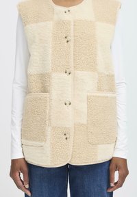 Bežs flīsa veste ar plecu dizainu, kurā ir četri kabatas, noapaļots kakls un piecas pogas priekšā.