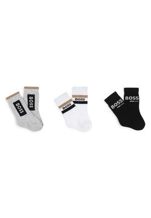 Trois paires de chaussettes en coton : grises avec une rayure marron, blanches avec une rayure noire et marron, et noires unies avec un logo blanc sur les bords.