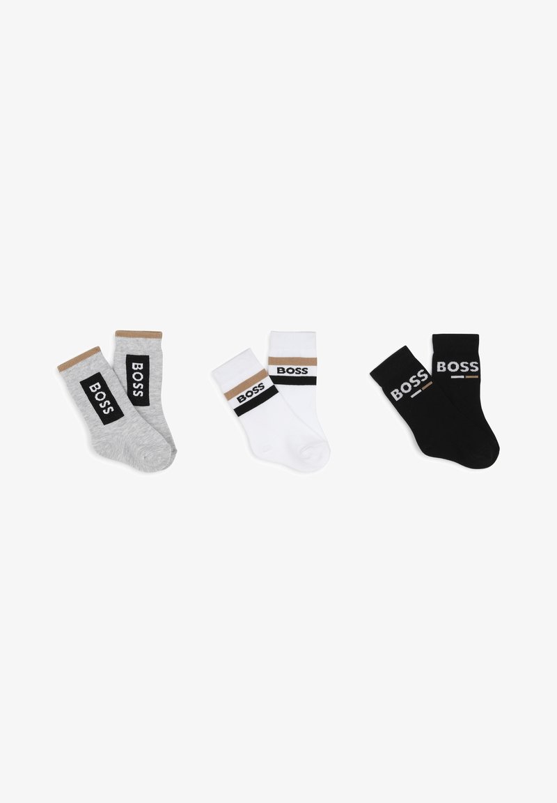Trois paires de chaussettes en coton : grises avec une rayure marron, blanches avec une rayure noire et marron, et noires unies avec un logo blanc sur les bords.