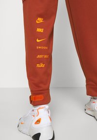 Oranga Nike-joggers har flera varumärkesloggor i gult, med en texturerad midjeband och justerbara muddar. Pairade med vita sneakers.