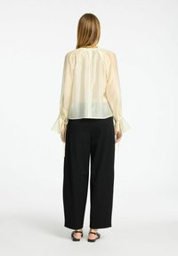 Blusa trasparente color crema con maniche lunghe e svasate, caratterizzata da dettagli a cravatta sui polsini, abbinata a pantaloni neri a vita alta e a gamba larga.