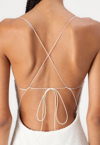 Πίσω όψη ενός φορέματος με λεπτές λευκές τιράντες που διασταυρώνονται στη χαμηλή πλάτη, αποκαλύπτοντας μια λεία υφή δέρματος και ένα υφασμάτινο σχέδιο σε ανοιχτό χρώμα.