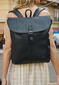 Zaino in pelle nera con chiusura a pattina, spallacci regolabili e dettaglio con fibbia frontale. La superficie ha una texture liscia e un design minimale.