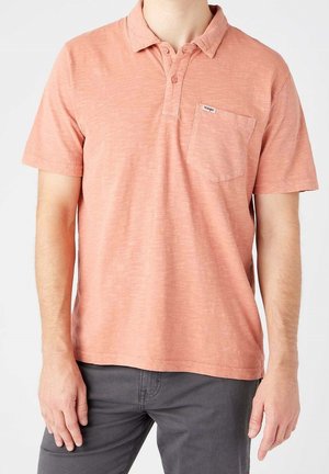 Homme portant un polo manches courtes de couleur saumon avec une poche poitrine et un pantalon gris foncé, debout devant un fond blanc.