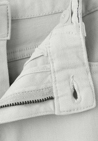 Ljusgrå denimtyg med detalj av dragkedja, med vit knappstängning och sömmar som accenter. Märkbart fransig kant nära dragkedjan.